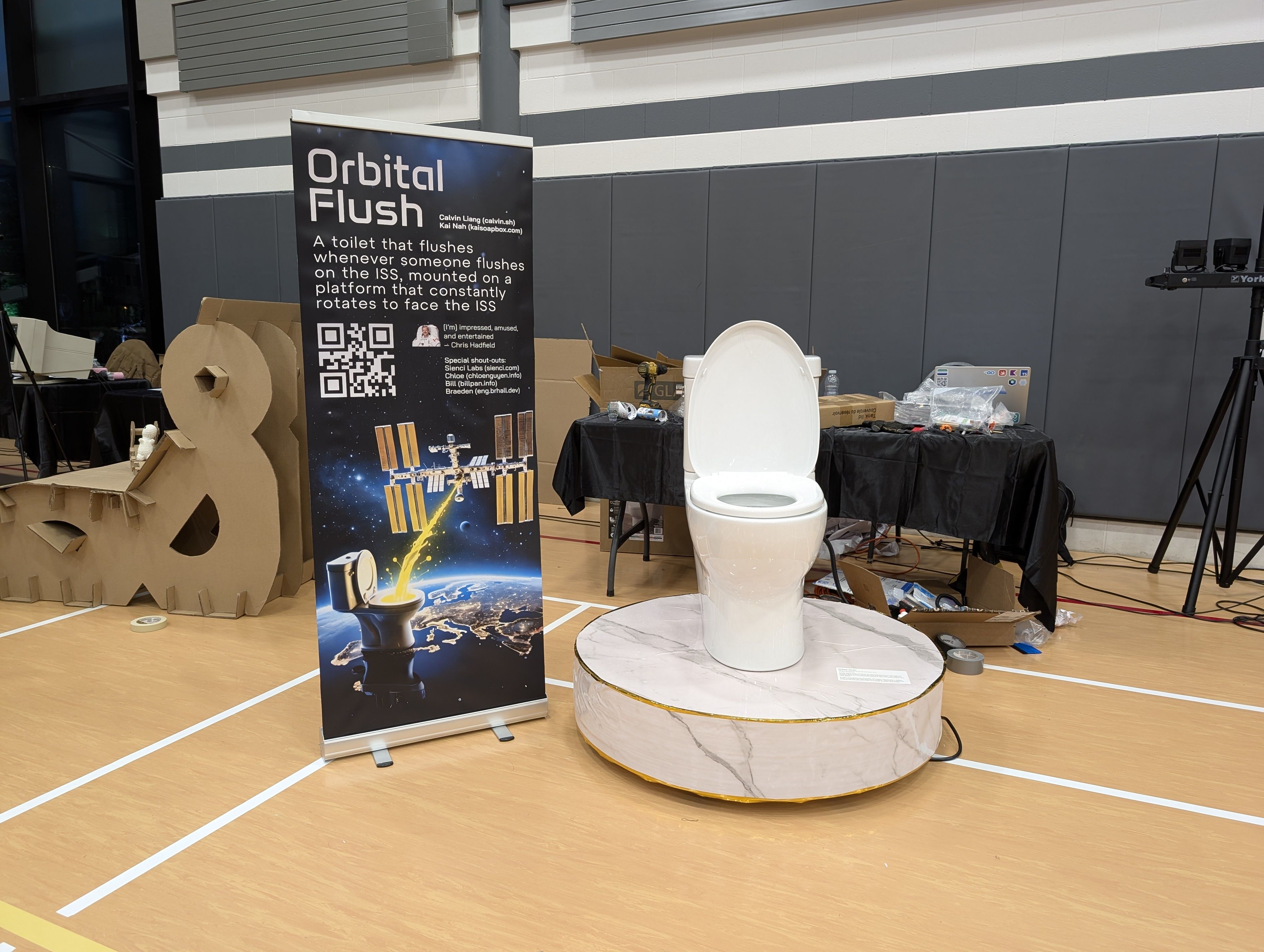 An art installation titled "Orbital Flush" displayed in a gymnasium, featuring a real toilet mounted on a circular platform that mimics marble. A nearby banner explains that the toilet flushes whenever someone flushes on the International Space Station, with illustrations showing a toilet shooting a stream toward the ISS. The setup includes tables with tools and supplies, cardboard sculptures, and a QR code on the banner.