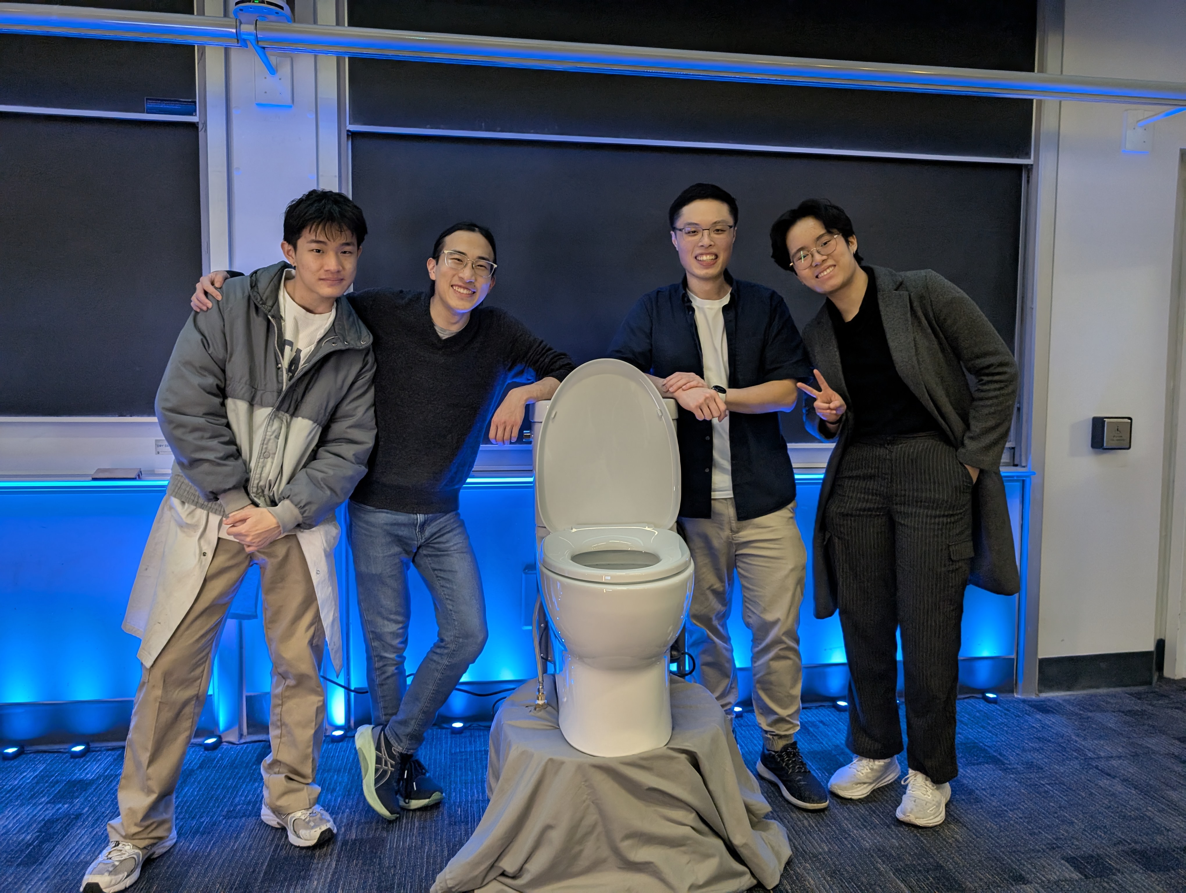 Four young adults: Bill, Kai, Calvin, and Chloe pose indoors in front of a toilet mounted on a small platform draped with gray fabric. They are smiling and casually dressed; Chloe, on the far right, is flashing a peace sign. The background features a classroom setting with large blackboards and blue accent lighting along the wall. The atmosphere is playful and lighthearted.