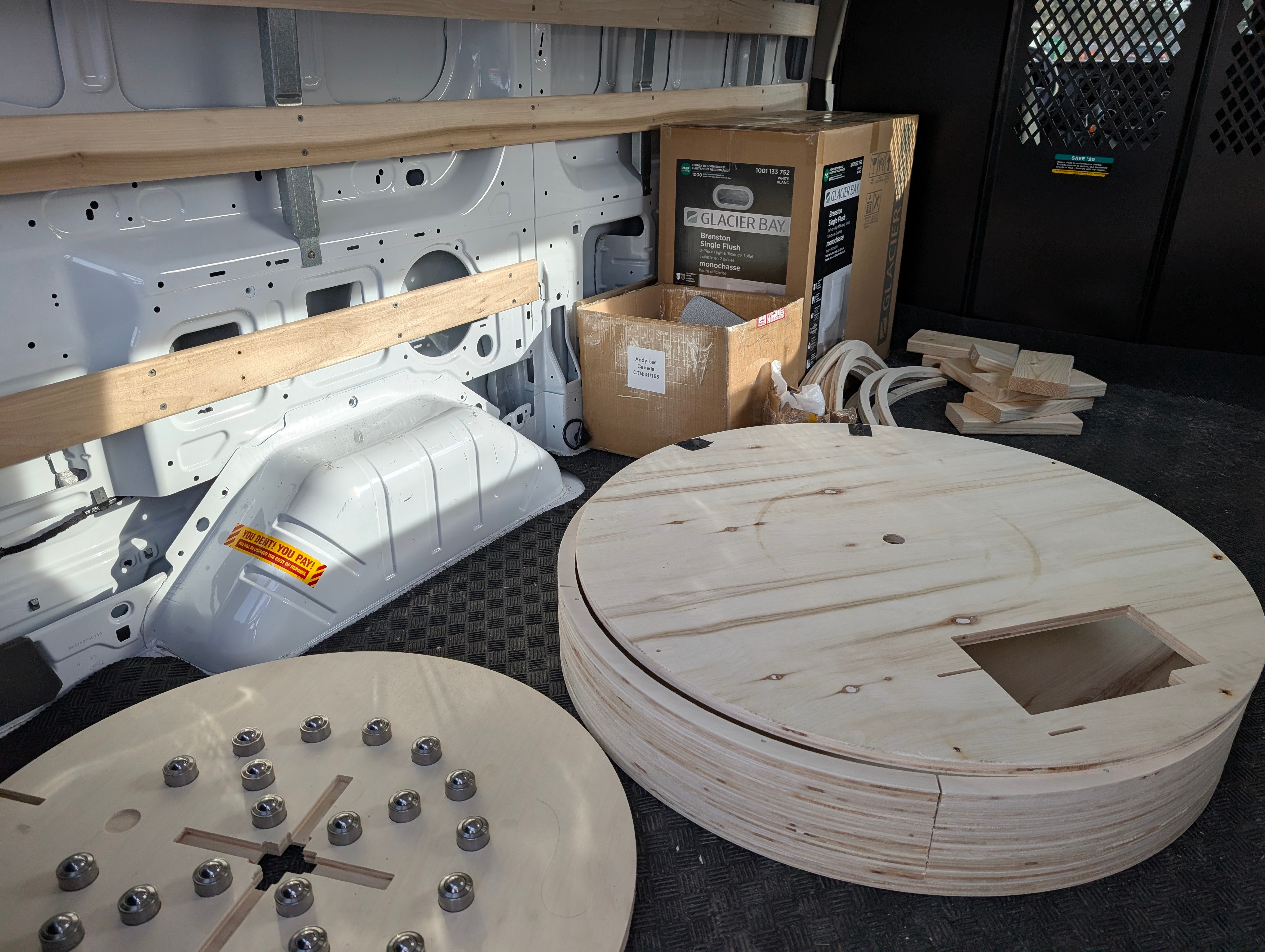Interior of a U-Haul van filled with woodworking materials, including large circular wooden discs, a Glacier Bay toilet box, curved wooden strips, cut lumber, and a partially assembled wooden piece with metal bearings.